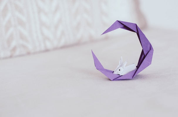 An origami sculpture featuring a small white rabbit resting within a crescent-shaped purple structure. The background is an off-white, textured surface that complements the delicate and intricate design of the origami.