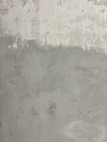 A textured concrete wall with subtle variations in tone and roughness. The surface shows patches of lighter and darker gray, with small visible pores and occasional faint markings.