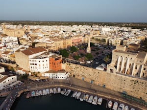 An aerial view of a coastal town with a historic Mediterranean atmosphere. The image features a harbor filled with small boats, bordered by a promenade. Behind the harbor, an assortment of colorful buildings and historic structures, including a prominent obelisk in a town square, extends into the distance. The architecture combines modern and classic elements, with terracotta roofs and beige stone facades.