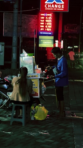 A nighttime street scene with a food vendor stall illuminated by neon lights in the background. A person is seated on a stool in front of the vendor, interacting with the seller. Various signs for currency exchange and money transfer are visible above.