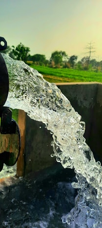 A stream of clear water is gushing from a pipe into a basin, with a backdrop of a lush green field and scattered trees under a clear sky.