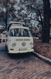 A vintage Volkswagen van is parked on a cobblestone street, surrounded by trees with dense foliage. The scene appears to be set in a park or outdoor area, with a relaxed and nostalgic ambience. The van is white and has a roof rack loaded with items, possibly camping equipment.