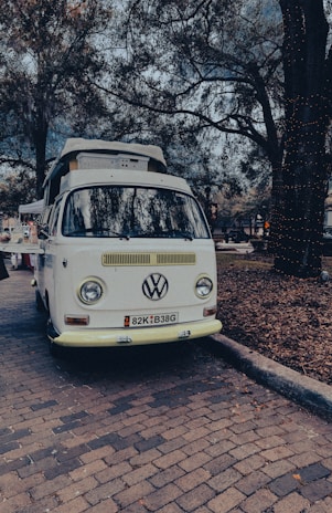 A vintage Volkswagen van is parked on a cobblestone street, surrounded by trees with dense foliage. The scene appears to be set in a park or outdoor area, with a relaxed and nostalgic ambience. The van is white and has a roof rack loaded with items, possibly camping equipment.