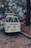 A vintage Volkswagen van is parked on a cobblestone street, surrounded by trees with dense foliage. The scene appears to be set in a park or outdoor area, with a relaxed and nostalgic ambience. The van is white and has a roof rack loaded with items, possibly camping equipment.