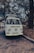 A vintage Volkswagen van is parked on a cobblestone street, surrounded by trees with dense foliage. The scene appears to be set in a park or outdoor area, with a relaxed and nostalgic ambience. The van is white and has a roof rack loaded with items, possibly camping equipment.