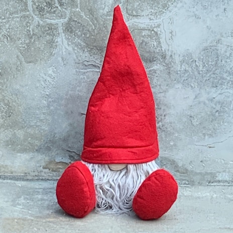 A small decorative gnome with a tall, pointed red hat and fluffy white beard sits against a textured, gray concrete wall. Its round nose peeks out from underneath the hat, and large, round red mittens rest on its side.