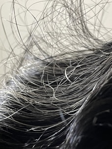 A close-up view of hair with numerous loose strands intertwined and overlapping. The strands appear to be gray or silver, indicating natural or dye-induced color. The background is blurred and neutral, focusing attention on the texture and flow of the hair.