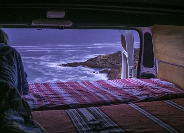 A cozy scene inside a van looking out onto a rocky coastline during dusk or dawn. The van door is open, revealing the ocean waves gently crashing against the rocks. The back of the van is furnished with a bed covered in red and white striped blankets, suggesting a relaxed camping setup. A person, partially visible, seems to be enjoying the view.