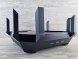 A sleek, black router with multiple angular antennas extending upwards and outwards on a wooden surface. The router has a modern design, featuring vents for airflow and buttons on the front panel. The background is a neutral, textured wall.