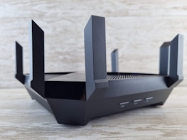 A sleek, black router with multiple angular antennas extending upwards and outwards on a wooden surface. The router has a modern design, featuring vents for airflow and buttons on the front panel. The background is a neutral, textured wall.