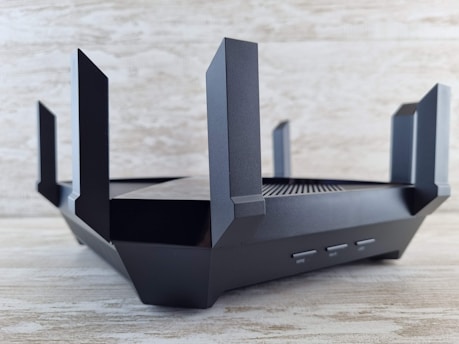 A sleek, black router with multiple angular antennas extending upwards and outwards on a wooden surface. The router has a modern design, featuring vents for airflow and buttons on the front panel. The background is a neutral, textured wall.