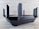 A sleek, black router with multiple angular antennas extending upwards and outwards on a wooden surface. The router has a modern design, featuring vents for airflow and buttons on the front panel. The background is a neutral, textured wall.