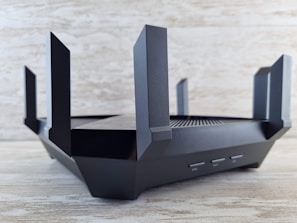 A sleek, black router with multiple angular antennas extending upwards and outwards on a wooden surface. The router has a modern design, featuring vents for airflow and buttons on the front panel. The background is a neutral, textured wall.