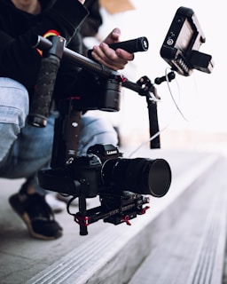 A camera setup attached to a stabilizer is being held. The equipment includes a camera with a large lens and various attachments, likely used for filming.