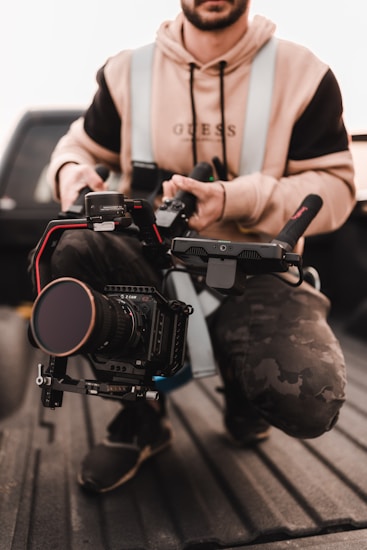 A person kneels in a vehicle holding a professional camera rig. The individual is wearing a casual outfit with a hoodie and a vest. The camera rig includes a large lens and is held securely, suggesting a focus on filming or photography.