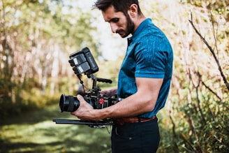 A person with a beard is holding and operating a professional video camera rig outdoors in a wooded area. They appear focused as they handle the equipment, which includes a camera, lenses, and various attachments. The background is lush with greenery and soft sunlight filters through the trees.