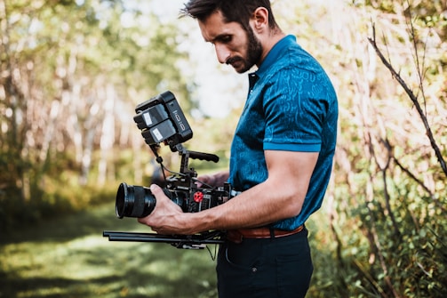 A person with a beard is holding and operating a professional video camera rig outdoors in a wooded area. They appear focused as they handle the equipment, which includes a camera, lenses, and various attachments. The background is lush with greenery and soft sunlight filters through the trees.