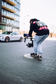 A person is bent over holding a camera stabilizer on a city street. They are wearing a black hoodie with colorful designs and light blue jeans. In the background, there is a modern high-rise building and a stationary silver car on a paved street.