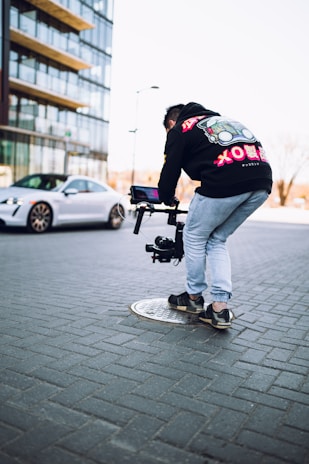A person is bent over holding a camera stabilizer on a city street. They are wearing a black hoodie with colorful designs and light blue jeans. In the background, there is a modern high-rise building and a stationary silver car on a paved street.