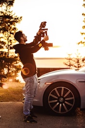 A person is standing next to a parked car, holding a professional camera setup, capturing images or video. The scene is bathed in warm, golden sunlight, suggesting an early morning or late afternoon setting. Trees are visible in the background, indicating an outdoor environment near a park or natural area.