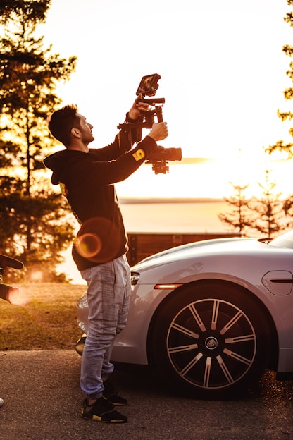 A person is standing next to a parked car, holding a professional camera setup, capturing images or video. The scene is bathed in warm, golden sunlight, suggesting an early morning or late afternoon setting. Trees are visible in the background, indicating an outdoor environment near a park or natural area.