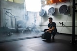 A man squats in a gym setting, surrounded by workout equipment such as a treadmill and some wall-mounted balls. He appears to be operating a smoke machine, as mist or smoke is billowing out around him. The environment suggests a fitness or training area with a somewhat moody and industrial atmosphere.