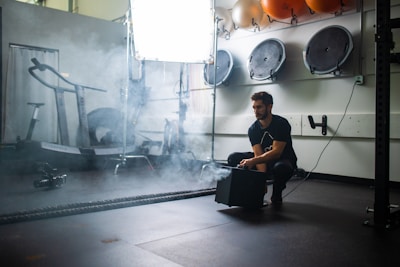 A man squats in a gym setting, surrounded by workout equipment such as a treadmill and some wall-mounted balls. He appears to be operating a smoke machine, as mist or smoke is billowing out around him. The environment suggests a fitness or training area with a somewhat moody and industrial atmosphere.