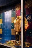 A mannequin is dressed in a beige and brown outfit, standing in a store display with a colorful pixelated backdrop made from multicolored blocks. The mannequin holds a purse and wears knee-high black boots. The background comprises a pattern of blue, red, yellow, and green colors, with a prominent display of the brand logo.