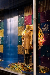 A mannequin is dressed in a beige and brown outfit, standing in a store display with a colorful pixelated backdrop made from multicolored blocks. The mannequin holds a purse and wears knee-high black boots. The background comprises a pattern of blue, red, yellow, and green colors, with a prominent display of the brand logo.