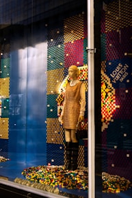 A mannequin is dressed in a beige and brown outfit, standing in a store display with a colorful pixelated backdrop made from multicolored blocks. The mannequin holds a purse and wears knee-high black boots. The background comprises a pattern of blue, red, yellow, and green colors, with a prominent display of the brand logo.