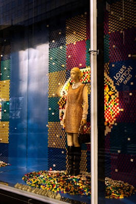 A mannequin is dressed in a beige and brown outfit, standing in a store display with a colorful pixelated backdrop made from multicolored blocks. The mannequin holds a purse and wears knee-high black boots. The background comprises a pattern of blue, red, yellow, and green colors, with a prominent display of the brand logo.