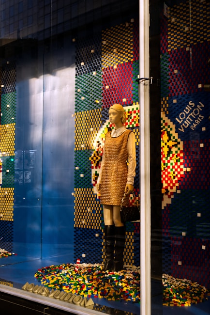 A mannequin is dressed in a beige and brown outfit, standing in a store display with a colorful pixelated backdrop made from multicolored blocks. The mannequin holds a purse and wears knee-high black boots. The background comprises a pattern of blue, red, yellow, and green colors, with a prominent display of the brand logo.