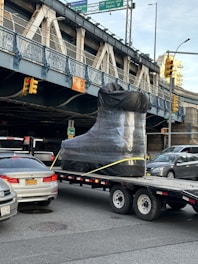 A large object shaped like a boot, wrapped in plastic, is being transported on a flatbed trailer down a city street. There are several cars around, including a BMW with a visible license plate. Overhead, there is a metal bridge with signage indicating Brooklyn, New York City, and a clearance warning of 12 feet. Traffic lights are visible, and the area seems busy with vehicles.