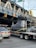 A large object shaped like a boot, wrapped in plastic, is being transported on a flatbed trailer down a city street. There are several cars around, including a BMW with a visible license plate. Overhead, there is a metal bridge with signage indicating Brooklyn, New York City, and a clearance warning of 12 feet. Traffic lights are visible, and the area seems busy with vehicles.