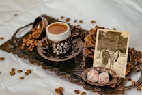 A vintage-style setup features a delicate cup of coffee on a decorative tray with intricately detailed borders. Scattered coffee beans add to the rustic aesthetic, alongside a sepia-toned photograph of people in an outdoor setting, evocative of past memories. Two pieces of Turkish delight rest in a small plate, enhancing the nostalgic feel.