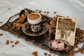 A vintage-style setup features a delicate cup of coffee on a decorative tray with intricately detailed borders. Scattered coffee beans add to the rustic aesthetic, alongside a sepia-toned photograph of people in an outdoor setting, evocative of past memories. Two pieces of Turkish delight rest in a small plate, enhancing the nostalgic feel.