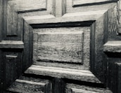 A detailed close-up of a wooden door with a geometric pattern. The wood appears to be dark and polished, showcasing its distinct grain and texture.