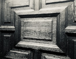 A detailed close-up of a wooden door with a geometric pattern. The wood appears to be dark and polished, showcasing its distinct grain and texture.