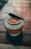 A cocktail garnished with a slice of dried citrus fruit and a whole dried pepper on top. The glass features a salted rim with red spices and is filled with ice.