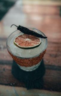 A cocktail garnished with a slice of dried citrus fruit and a whole dried pepper on top. The glass features a salted rim with red spices and is filled with ice.
