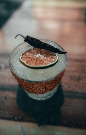 A cocktail garnished with a slice of dried citrus fruit and a whole dried pepper on top. The glass features a salted rim with red spices and is filled with ice.