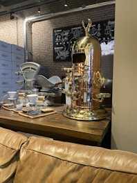 A vintage-style coffee machine with a polished copper finish stands prominently on a wooden table. Nearby, there are coffee brewing equipment like drippers and carafes arranged neatly. The background features brick walls and metallic pipes, giving an industrial vibe. A chart related to coffee is mounted on the wall.