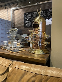 A vintage-style coffee machine with a polished copper finish stands prominently on a wooden table. Nearby, there are coffee brewing equipment like drippers and carafes arranged neatly. The background features brick walls and metallic pipes, giving an industrial vibe. A chart related to coffee is mounted on the wall.