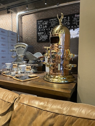 A vintage-style coffee machine with a polished copper finish stands prominently on a wooden table. Nearby, there are coffee brewing equipment like drippers and carafes arranged neatly. The background features brick walls and metallic pipes, giving an industrial vibe. A chart related to coffee is mounted on the wall.
