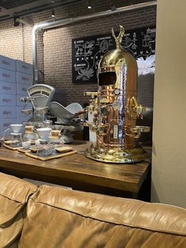 A vintage-style coffee machine with a polished copper finish stands prominently on a wooden table. Nearby, there are coffee brewing equipment like drippers and carafes arranged neatly. The background features brick walls and metallic pipes, giving an industrial vibe. A chart related to coffee is mounted on the wall.
