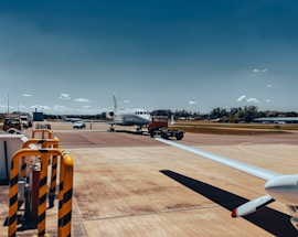 A small private jet is positioned on an airport tarmac with a clear blue sky in the background. The aircraft appears to be undergoing maintenance or preparation, as a red tow vehicle is attached to it at the front. Surrounding the plane are various airport vehicles and equipment. The scene includes a wide expanse of the runway and grassy areas in the distance.