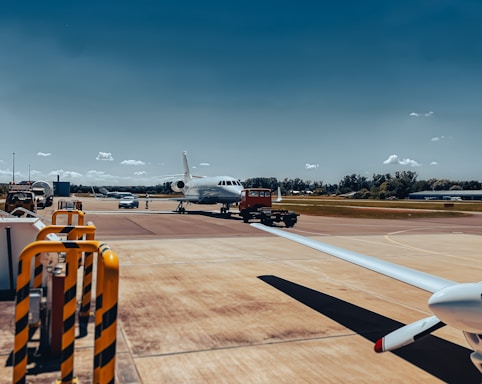 A small private jet is positioned on an airport tarmac with a clear blue sky in the background. The aircraft appears to be undergoing maintenance or preparation, as a red tow vehicle is attached to it at the front. Surrounding the plane are various airport vehicles and equipment. The scene includes a wide expanse of the runway and grassy areas in the distance.