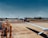 A small private jet is positioned on an airport tarmac with a clear blue sky in the background. The aircraft appears to be undergoing maintenance or preparation, as a red tow vehicle is attached to it at the front. Surrounding the plane are various airport vehicles and equipment. The scene includes a wide expanse of the runway and grassy areas in the distance.