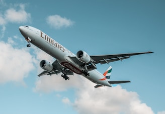 A large Emirates airplane soars through the sky with the landing gear retracted. The aircraft is prominently branded with the Emirates logo and features a white body with red, green, and black markings on the tail. The sky is mostly clear with a few scattered clouds.