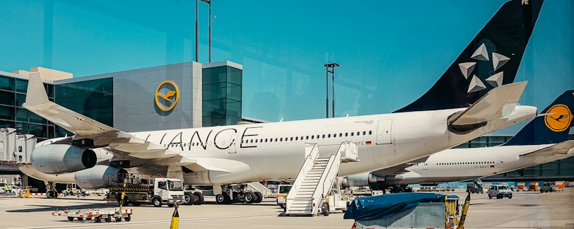 An airplane is parked on an airport tarmac, with its engines and landing gear visible. The airplane has a white body with a logo on its tail fin. An aircraft boarding stair is positioned next to the plane, and there are small vehicles and equipment scattered around, suggesting ground operations. The backdrop includes an airport terminal building with clear skies above.